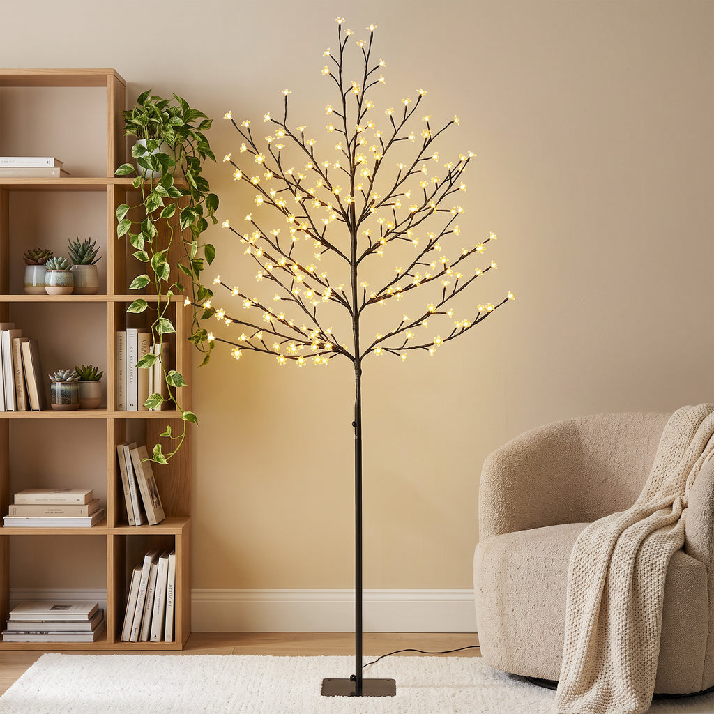 Árbol de cerezo luminoso con luz blanca cálida junto a una estantería de madera y un sillón