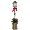 Farol de Navidad LED con linterna de bronce, lazo rojo y pequeño árbol verde en una maceta, vista frontal sobre fondo blanco