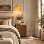 Lámpara de pie en arco naranja junto a una cama con mesilla de madera, planta y ropa de cama beige