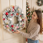 Mujer colgando la corona de Navidad nevada en una puerta junto a un árbol de Navidad