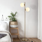 Lámpara de pie dorada con dos globos de cristal junto a una cama y una planta en un dormitorio