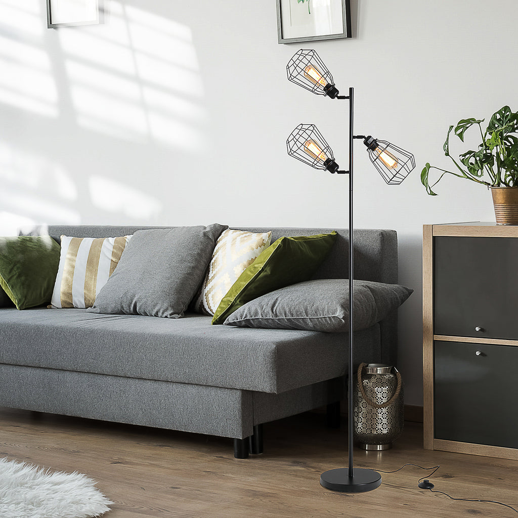 Lámpara de pie industrial de tres luces encendida junto a un sofá gris y una planta