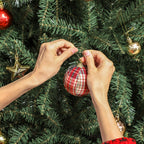 Manos colocando una bola de Navidad a cuadros en las ramas del abeto