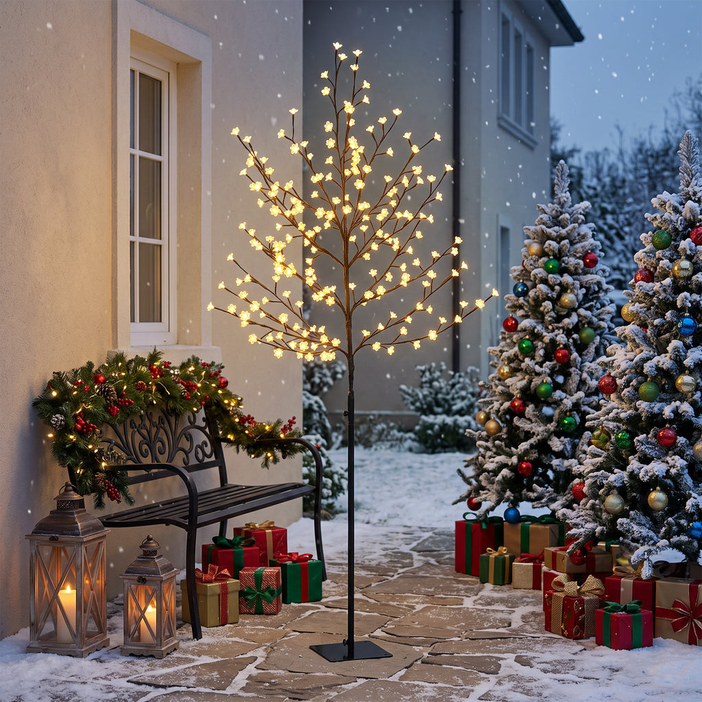 Árbol de cerezo luminoso con luz cálida junto a árboles de Navidad y regalos en la entrada de una casa