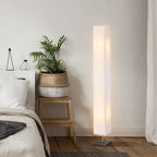 Lámpara de pie de columna encendida junto a una cama, una cesta y una planta