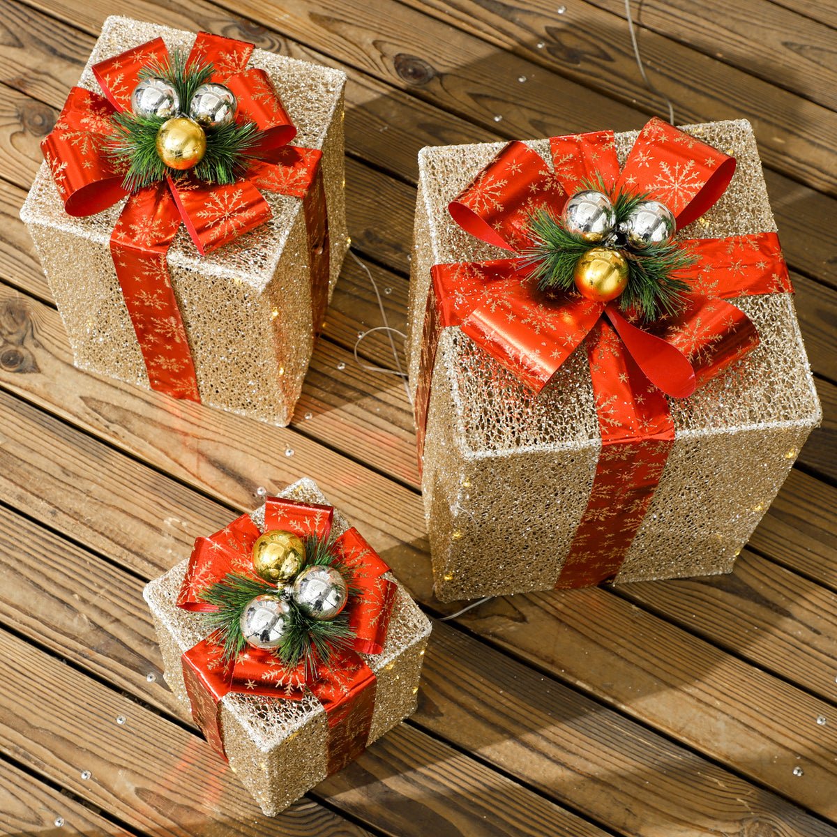 Tres cajas de regalo doradas con lazo rojo y bolas plateadas y doradas sobre una terraza de madera